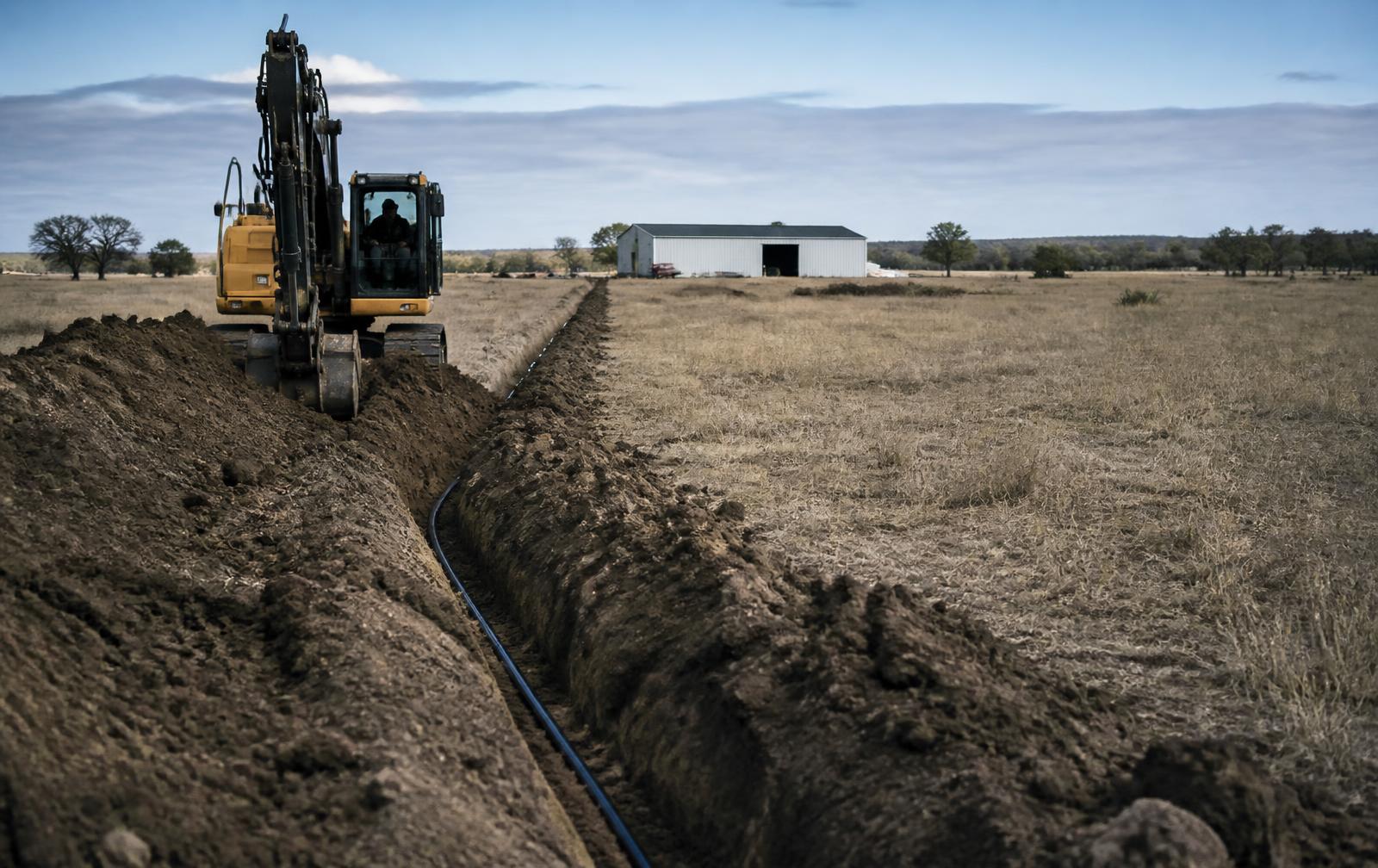 Excavator trenching a utility line across open ranch land toward a metal outbuilding